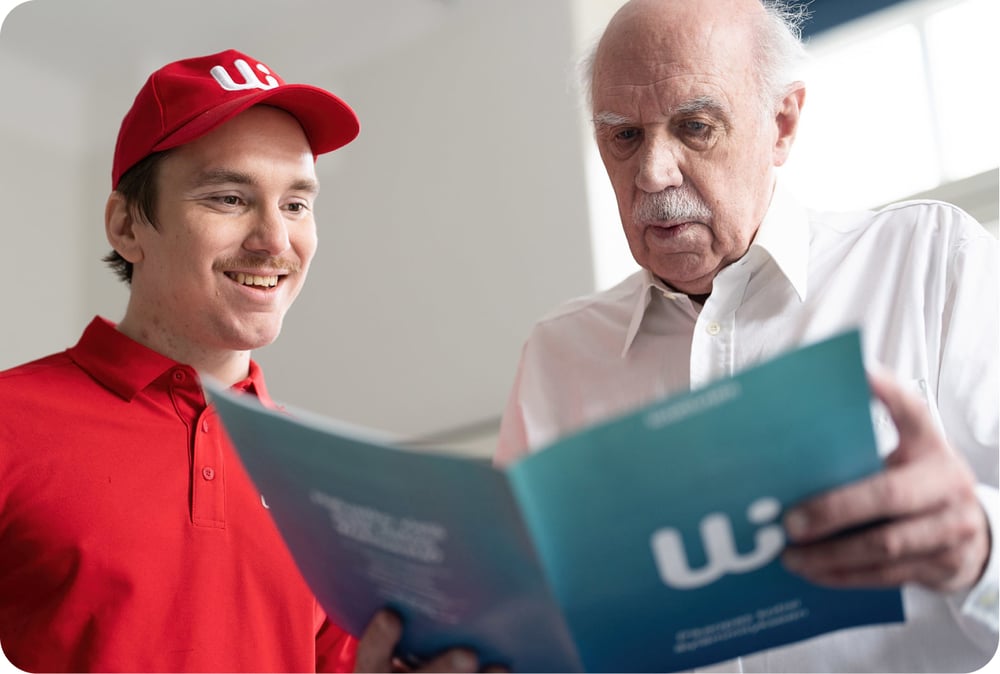 Vanha mies lukee Wattisen opasta ja Wattisen edustaja punaisessa paidassa hymyilee vieressä. 