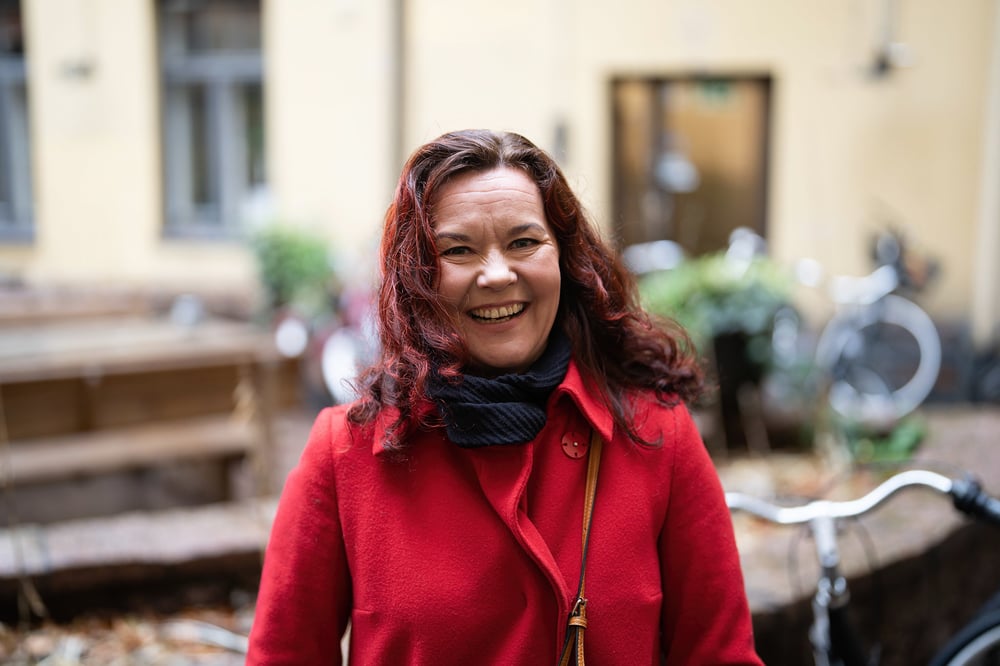 Punatakkinen nainen seisoo talon pihalla ja hymyilee kohti.